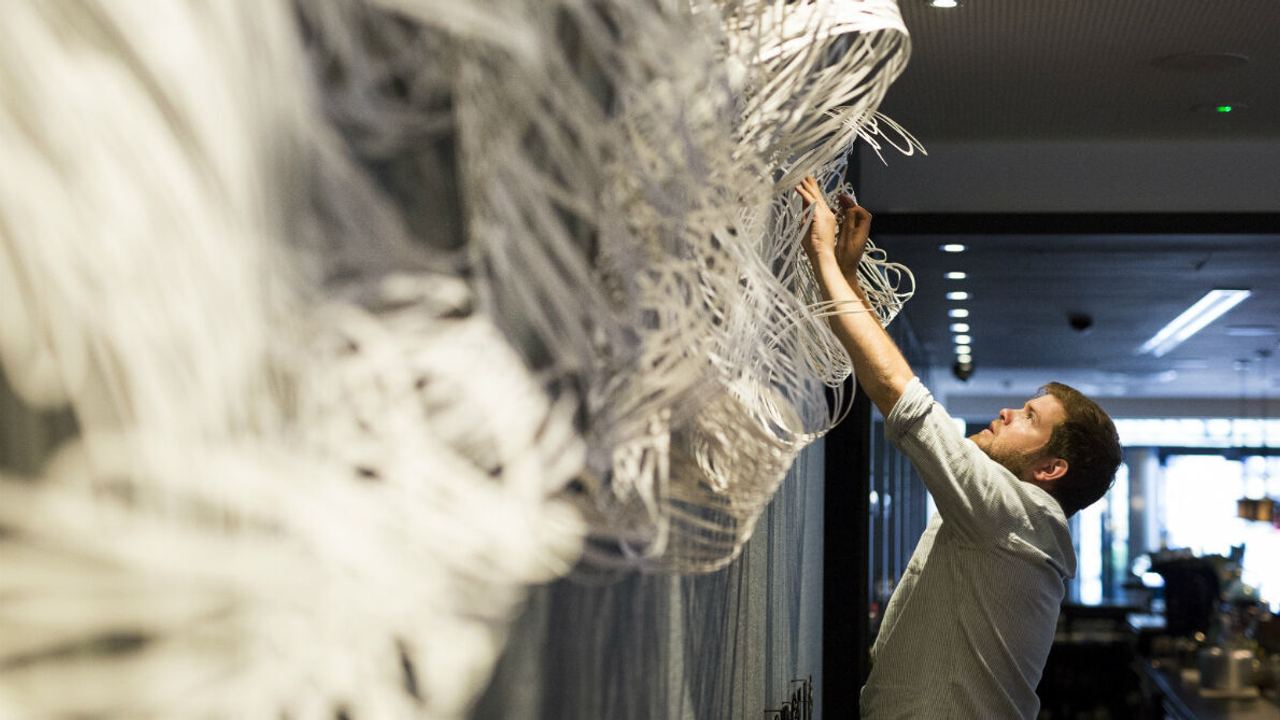 Ein Mann arrangiert oder repariert eine große, komplexe, weiße Wandinstallation in einem modernen Innenraum mit gedämpfter Beleuchtung.