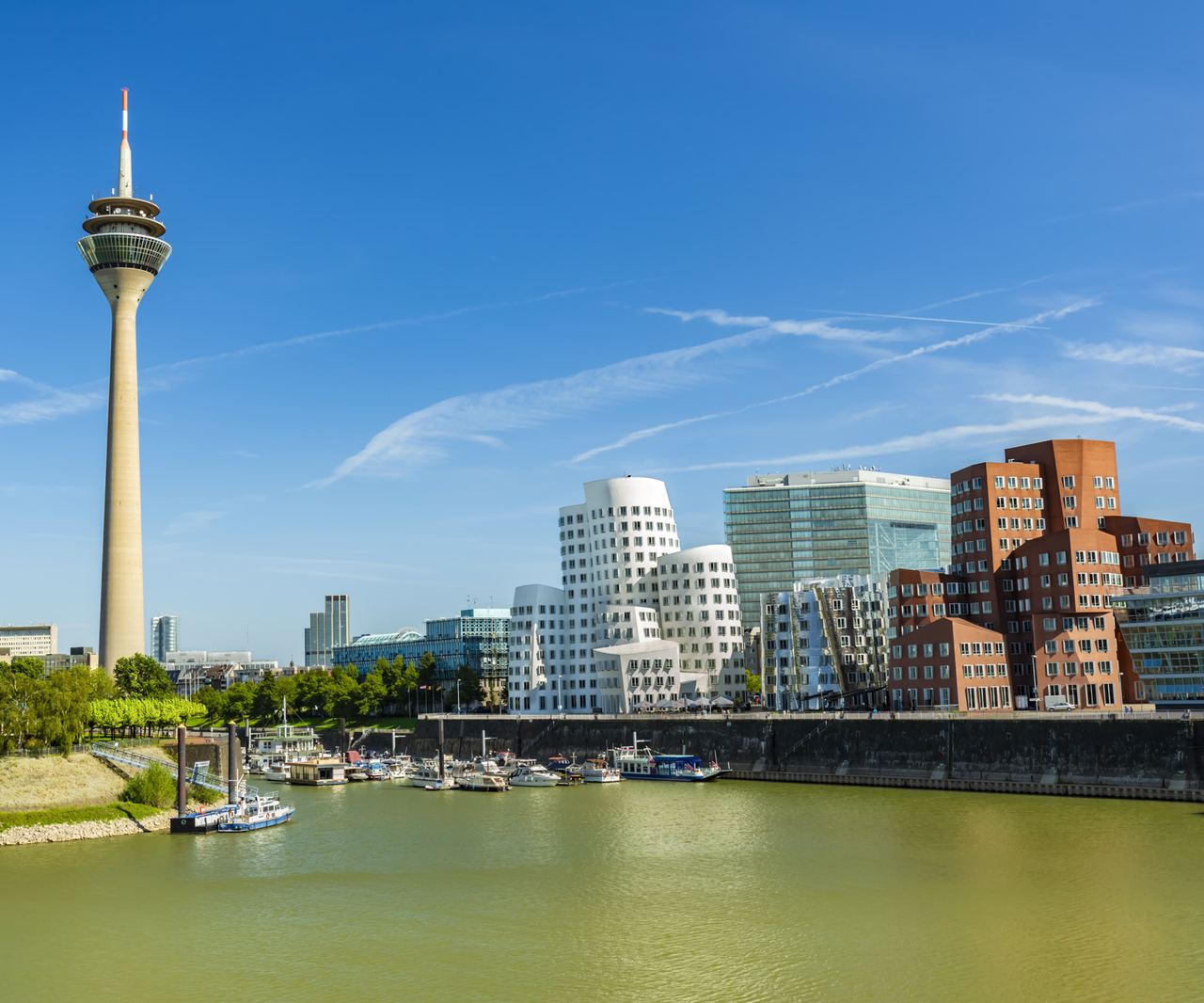 Panoramablick auf den Rhein mit der Düsseldorfer Skyline, inklusive des markanten Rheinturms und moderner, architektonisch auffälliger Gebäude am Ufer bei klarem Himmel. Panoramablick auf den Rhein mit der Düsseldorfer Skyline, inklusive des markanten Rheinturms und moderner, architektonisch auffälliger Gebäude am Ufer bei klarem Himmel.