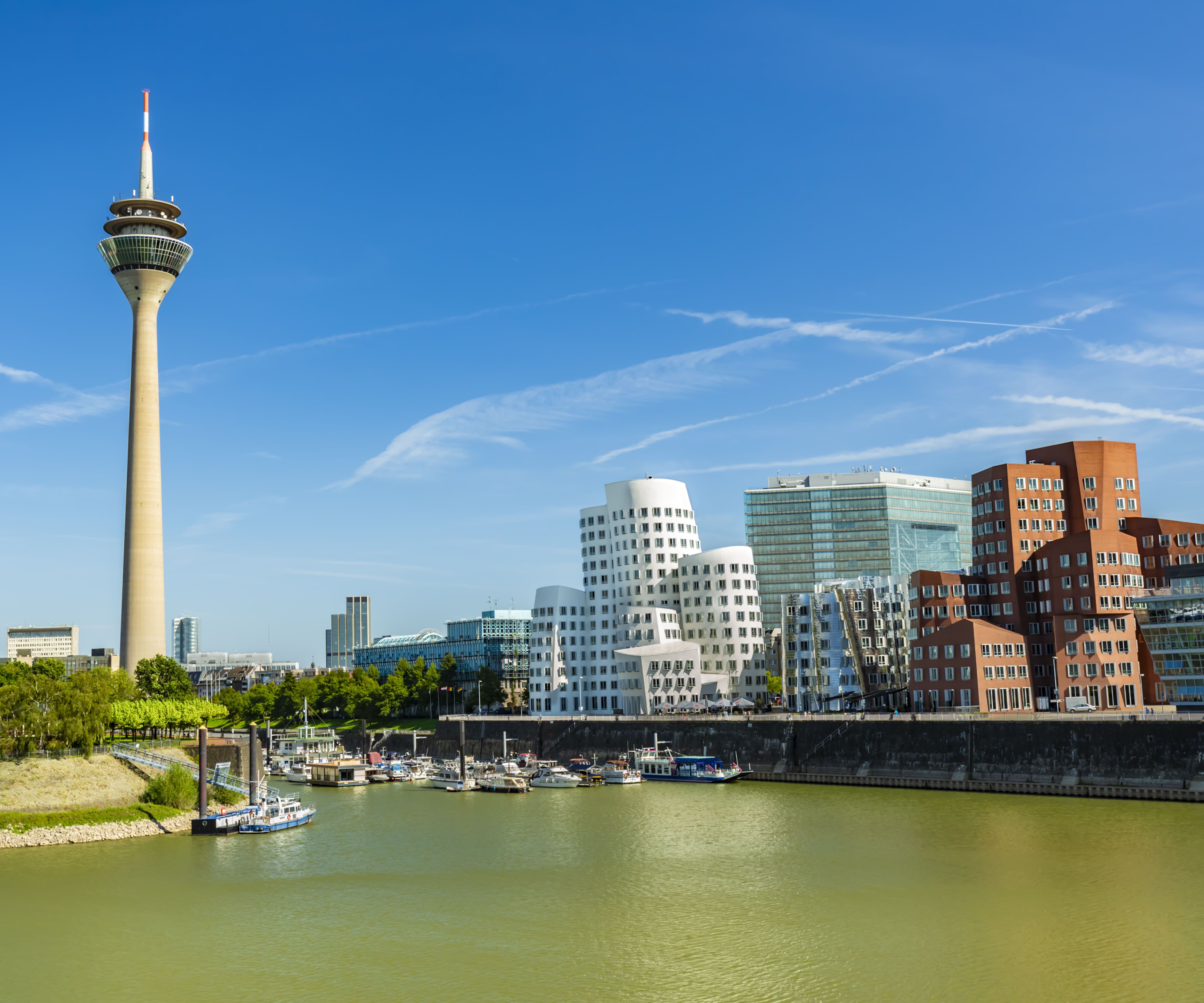 Panoramablick auf den Rhein mit der Düsseldorfer Skyline, inklusive des markanten Rheinturms und moderner, architektonisch auffälliger Gebäude am Ufer bei klarem Himmel.