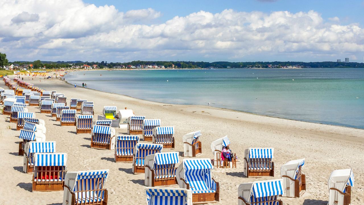 Strand mit vielen blau-weiß gestreiften Strandkörben, ruhiges Meer und teilweise bewölkter Himmel, ideal für erholsame Tage am Wasser.
