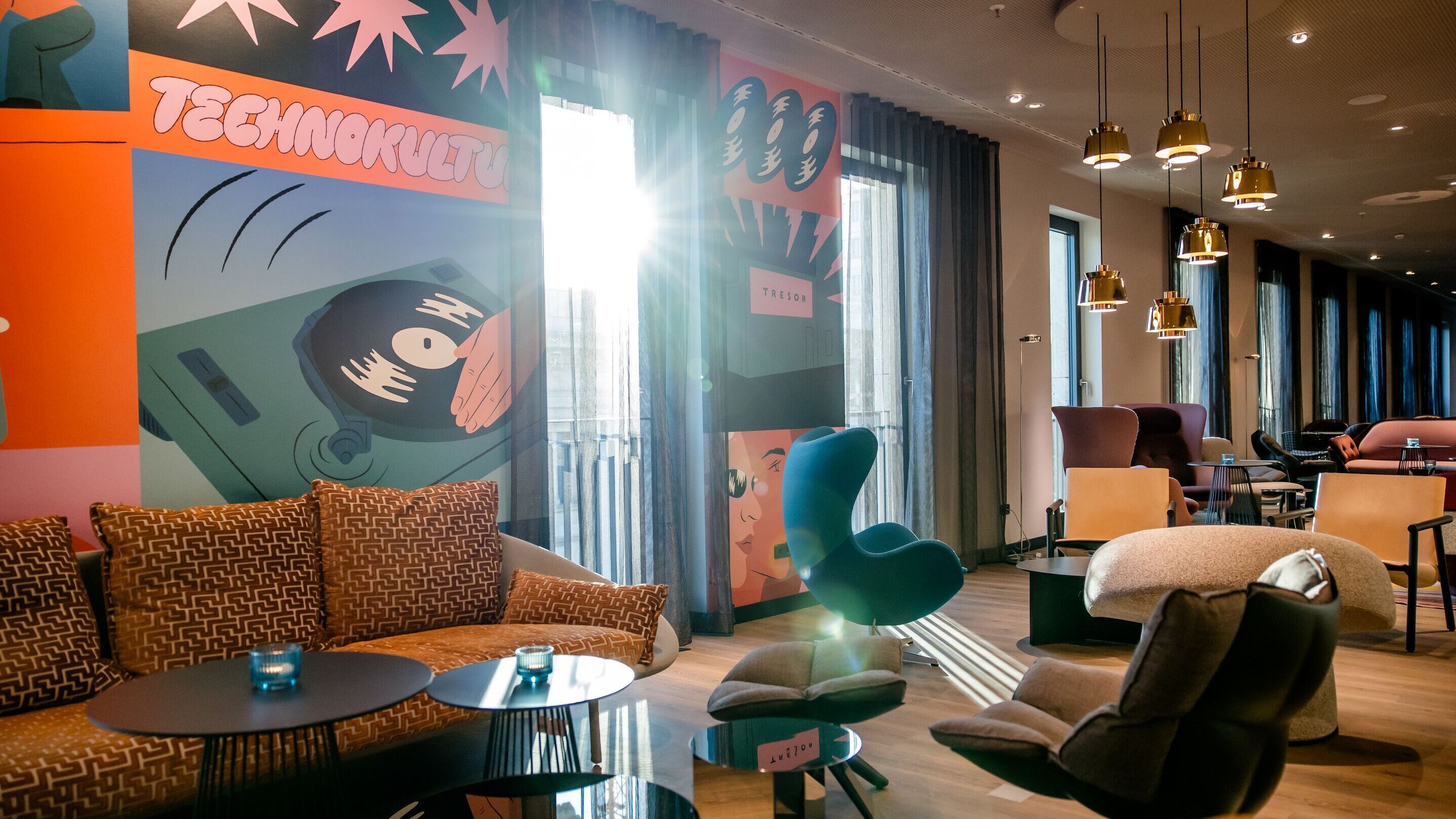 Modernes Lounge-Ambiente mit stilvollen Sesseln, Sofas und warmem Tageslicht. Farbige Wanddekoration mit Retro-Design sorgt für lebendige Atmosphäre, ideal zum Entspannen oder geselligen Beisammensein.