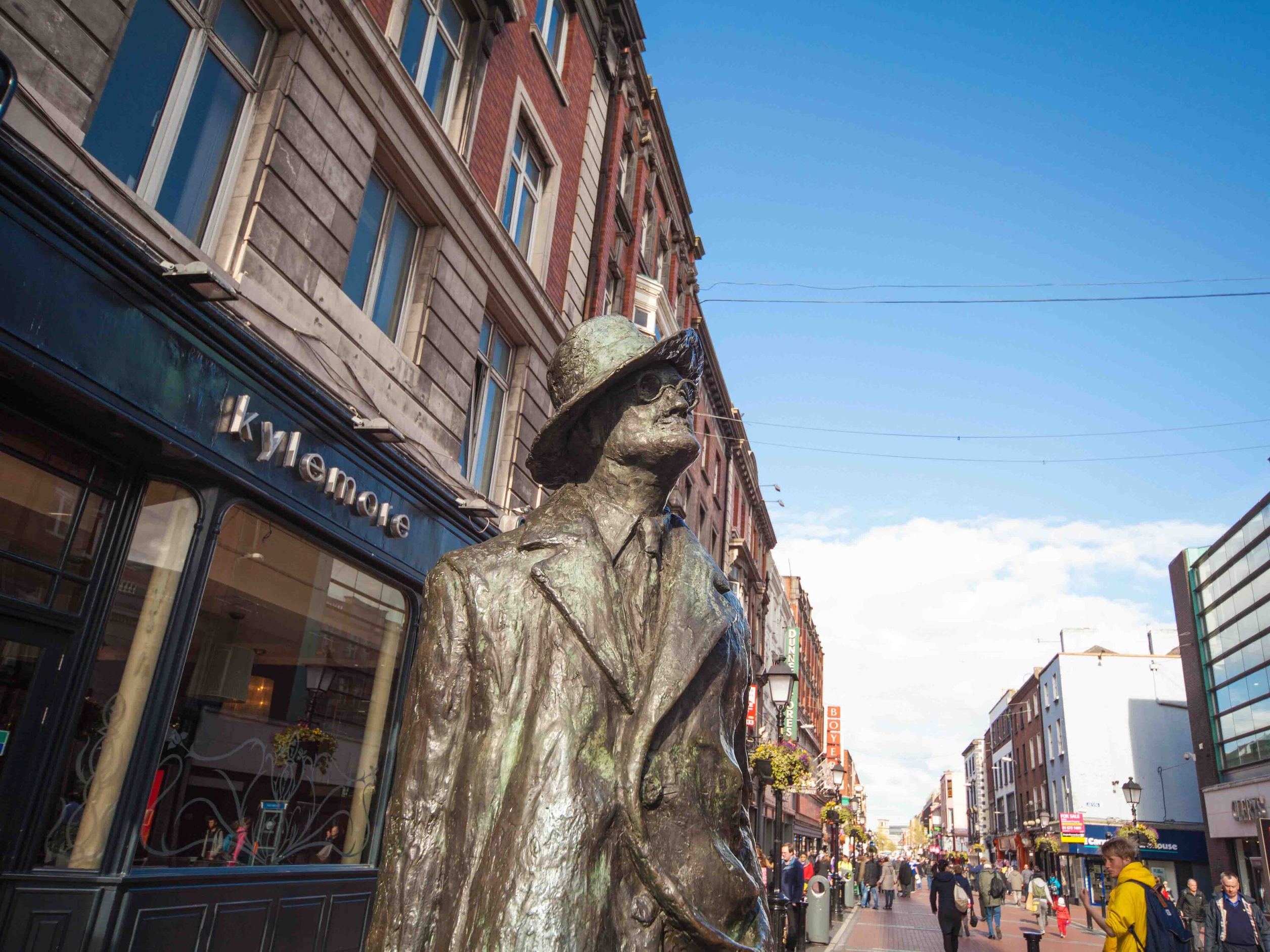 Bronzestatue eines Mannes mit Hut und Mantel auf einer belebten Einkaufsstraße unter klarem Himmel.