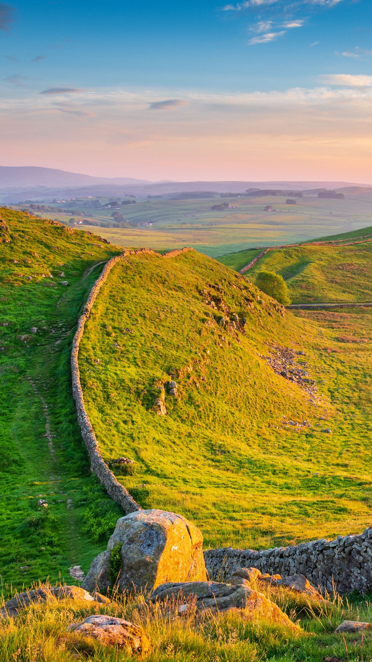 Weite grüne Hügel im Abendlicht, durchzogen von einer historischen, aus Natursteinen gefertigten Mauer, die sich entlang der Landschaft schlängelt. Ruhige, ländliche Atmosphäre mit weitem Blick in den sanft gebogenen Talraum unter einem farbenfrohen Himmel.