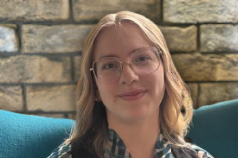 Junge Frau mit Brille lächelt freundlich, trägt eine gemusterte Bluse und schwarzen Ärmellosen Überwurf, sitzt vor einer Steinwand auf einem blauen Sessel. Junge Frau mit Brille lächelt freundlich, trägt eine gemusterte Bluse und schwarzen Ärmellosen Überwurf, sitzt vor einer Steinwand auf einem blauen Sessel.
