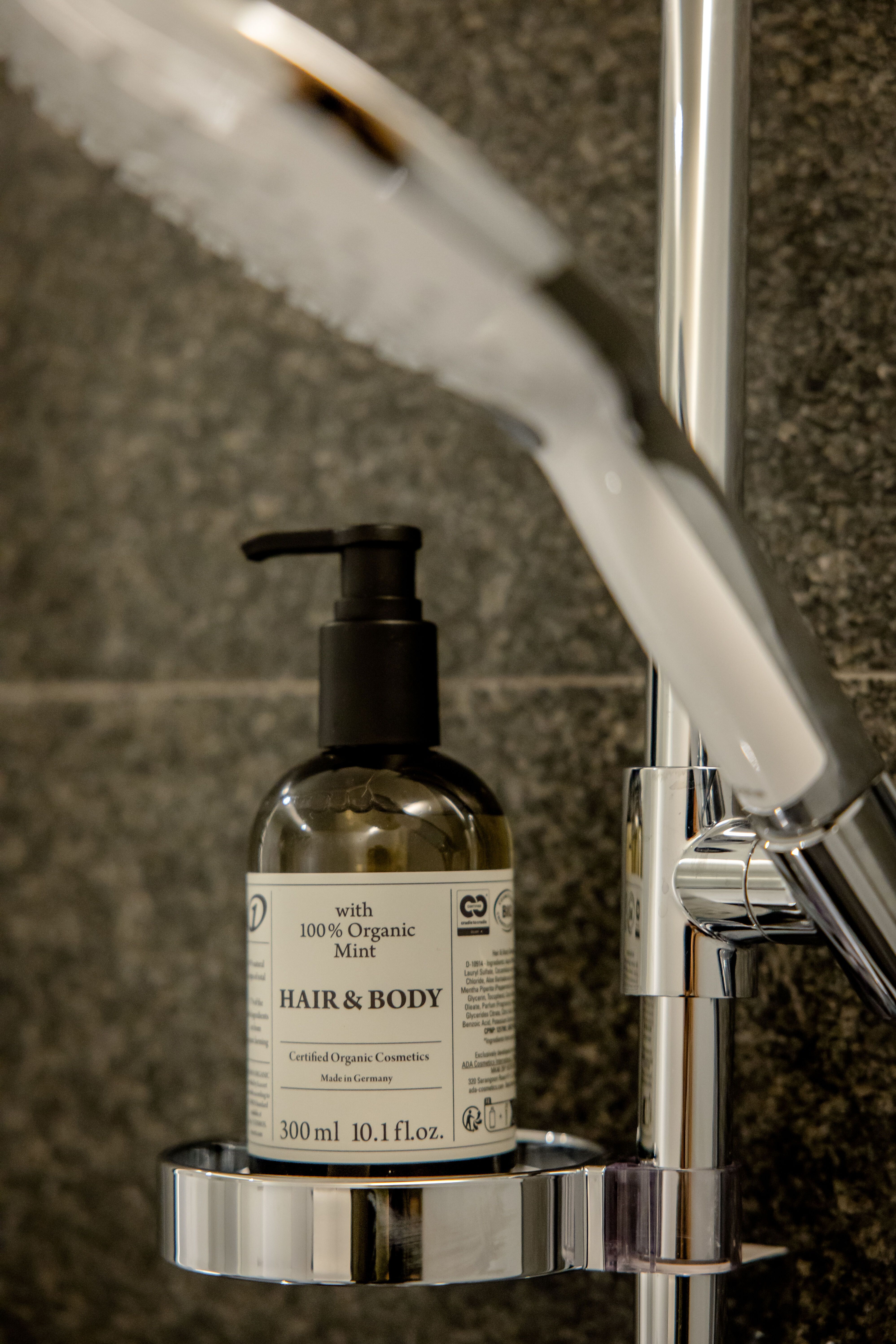 Flasche mit biologischem Haar- und Körperwaschmittel mit Minzduft, platziert in einem chromglänzenden Halter neben einer modernen Duschbrause vor dunklem Fliesenhintergrund.