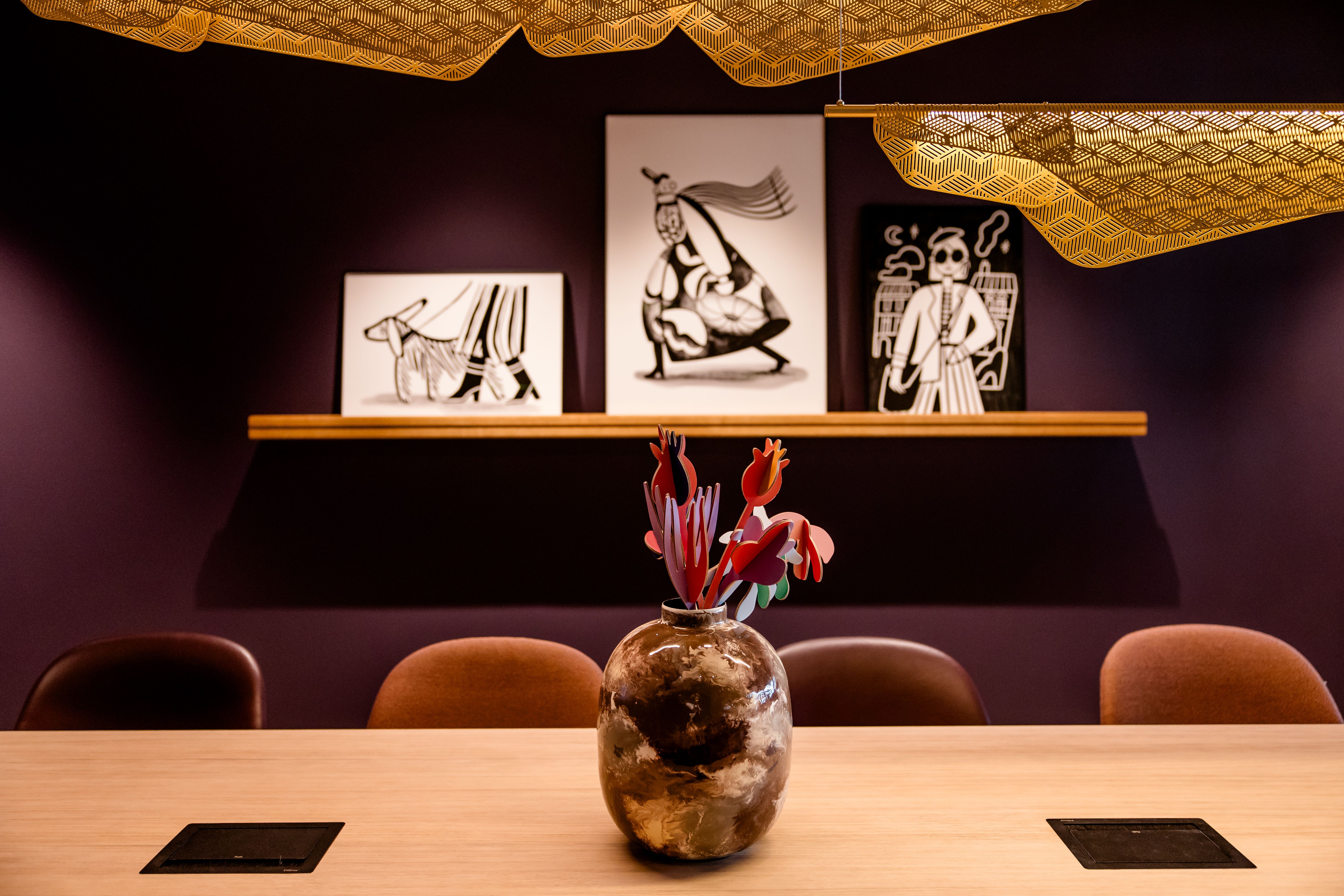 Ein stilvoll eingerichteter Besprechungsraum mit dunkler Wand, moderner Blumenvase auf dem Holztisch und drei schwarz-weißen Kunstwerken an der Wand, die abstrakte Figuren zeigen. Dezente Beleuchtung und elegante Akzente schaffen eine entspannte Atmosphäre.