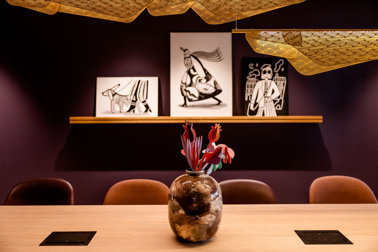 Ein stilvoll eingerichteter Besprechungsraum mit dunkler Wand, moderner Blumenvase auf dem Holztisch und drei schwarz-weißen Kunstwerken an der Wand, die abstrakte Figuren zeigen. Dezente Beleuchtung und elegante Akzente schaffen eine entspannte Atmosphäre.