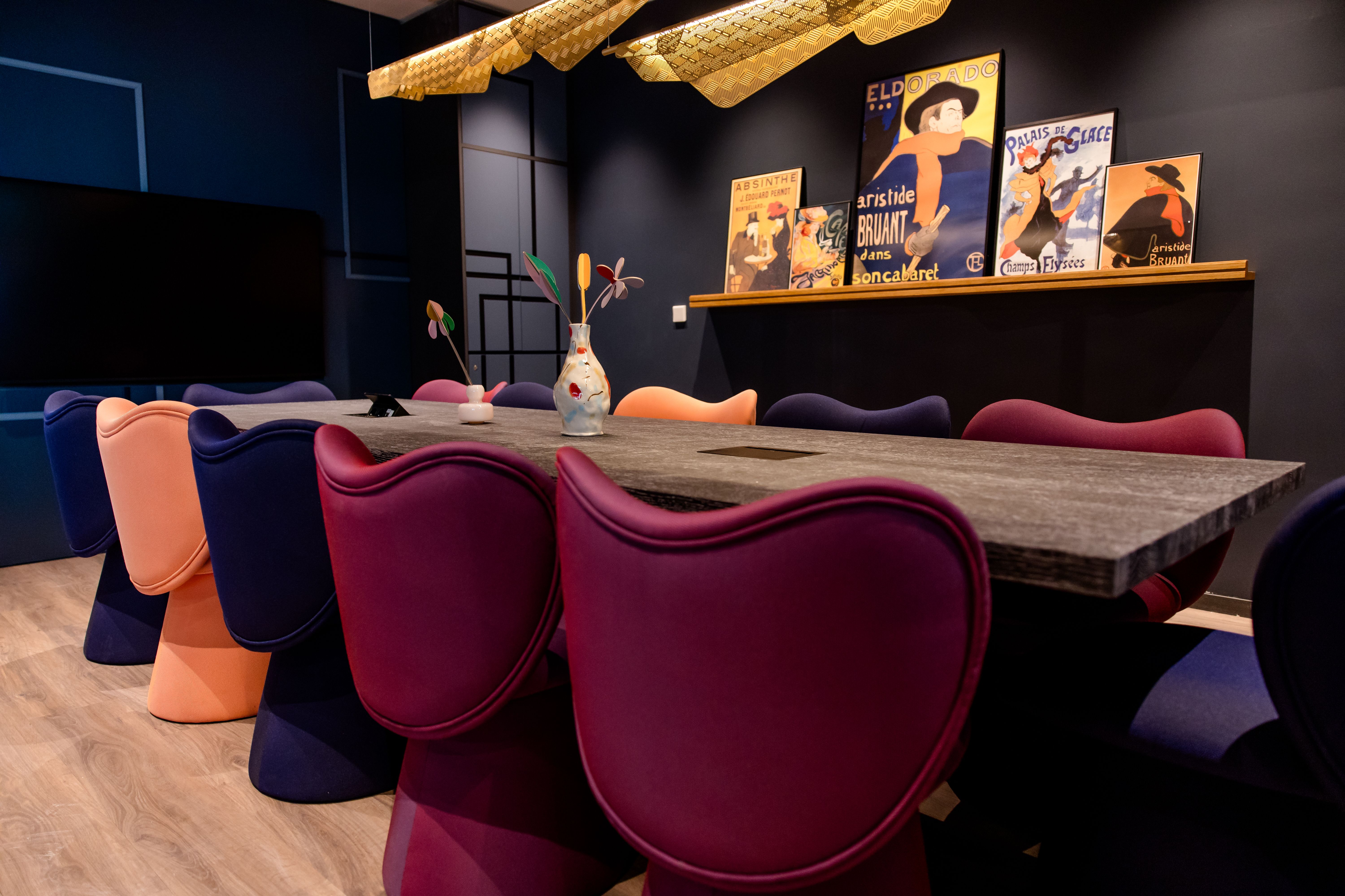 Moderner Besprechungsraum mit langem Tisch, bunten gepolsterten Stühlen und stilvollen vintage Plakaten an der Wand, warme Beleuchtung schafft ein einladendes Ambiente für Meetings oder gemeinsames Arbeiten.