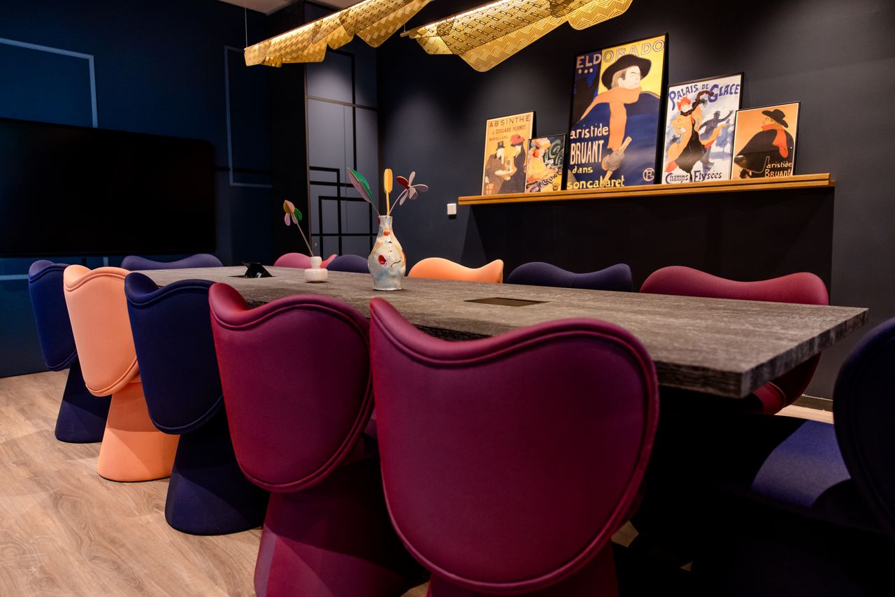 Moderner Besprechungsraum mit langem Tisch, bunten gepolsterten Stühlen und stilvollen vintage Plakaten an der Wand, warme Beleuchtung schafft ein einladendes Ambiente für Meetings oder gemeinsames Arbeiten.