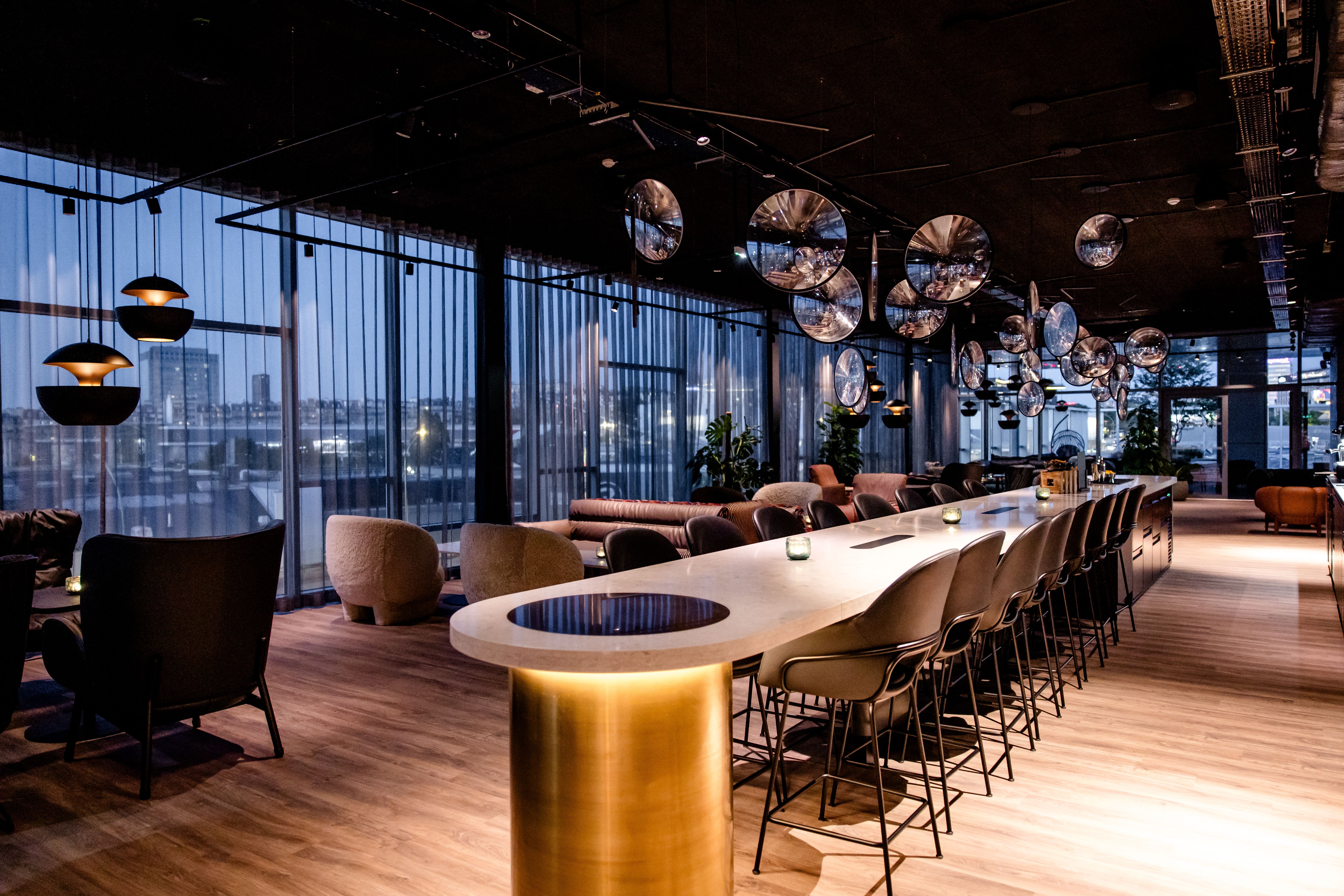 Moderne Hotelbar mit elegant beleuchtetem Tresen, stilvollen Sitzgelegenheiten und großflächigen Fenstern mit Ausblick auf die Stadt, atmosphärisch und einladend gestaltet für entspannte Abende und geselliges Beisammensein.