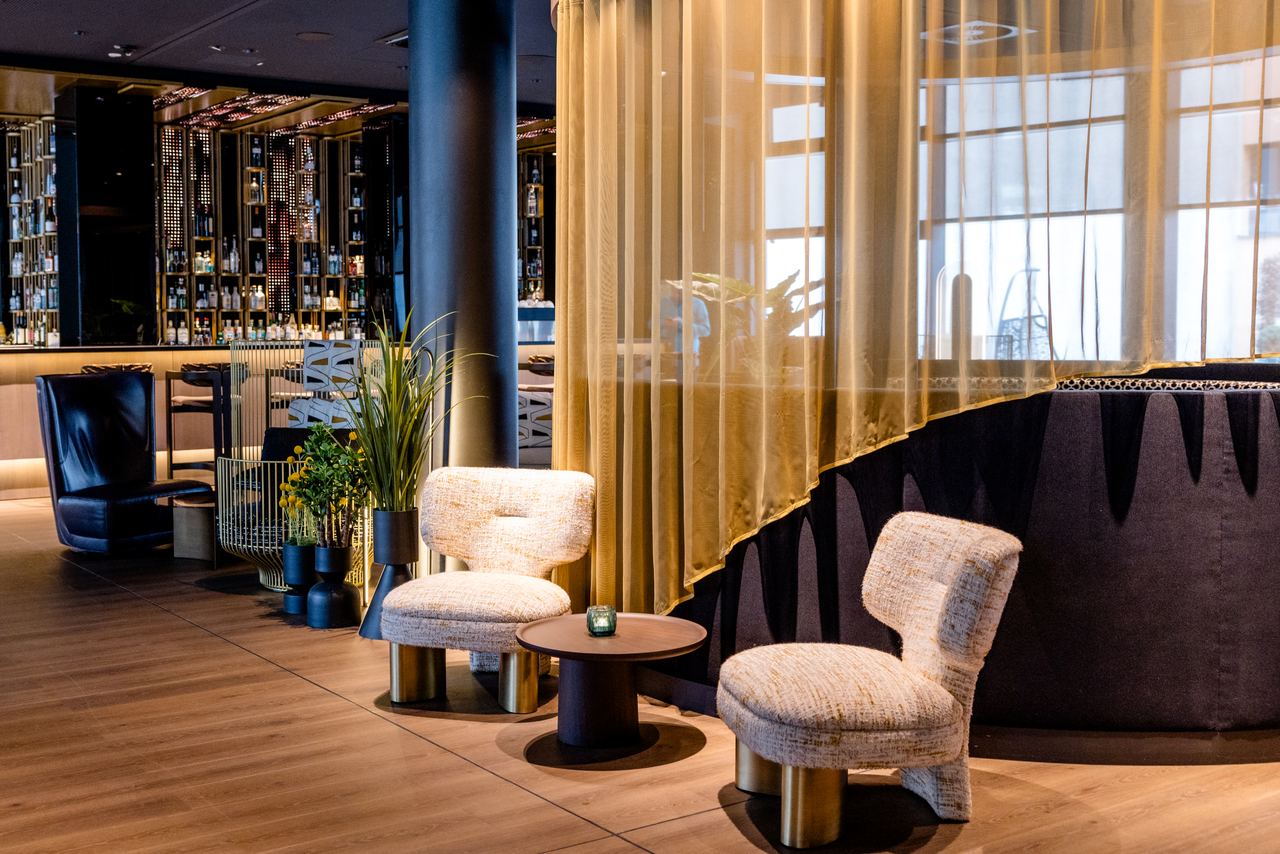 Eleganter Hotelbarbereich mit gemütlichen, modernen Sesseln, kleinem Beistelltisch und warmem, einladendem Licht. Transparenter Vorhang trennt den Raum dezent ab, schafft ruhige Atmosphäre zum Entspannen und Genießen.